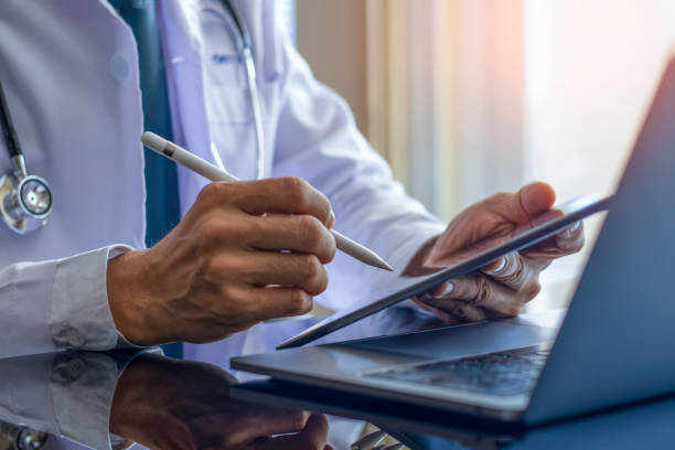 Male doctor in white coat with medical stethoscope, hand holding and using modern digital tablet, work on laptop computer at office. Telehealth, Online medical networking concept.