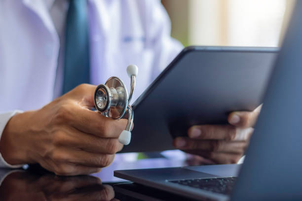 Male doctor in white lab coat, hand holding medical stethoscope and using modern digital tablet pc,
work on laptop computer at workplace. Online medical, e health or medical network concept.
